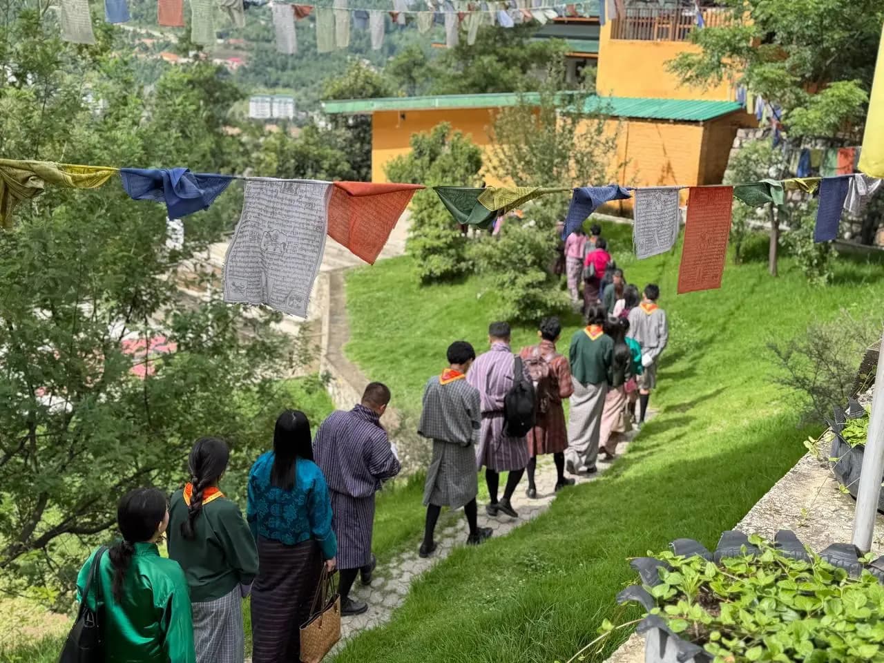 ELC students and teachers walking single file along a grass path under prayer flags, the Thimphu valley below