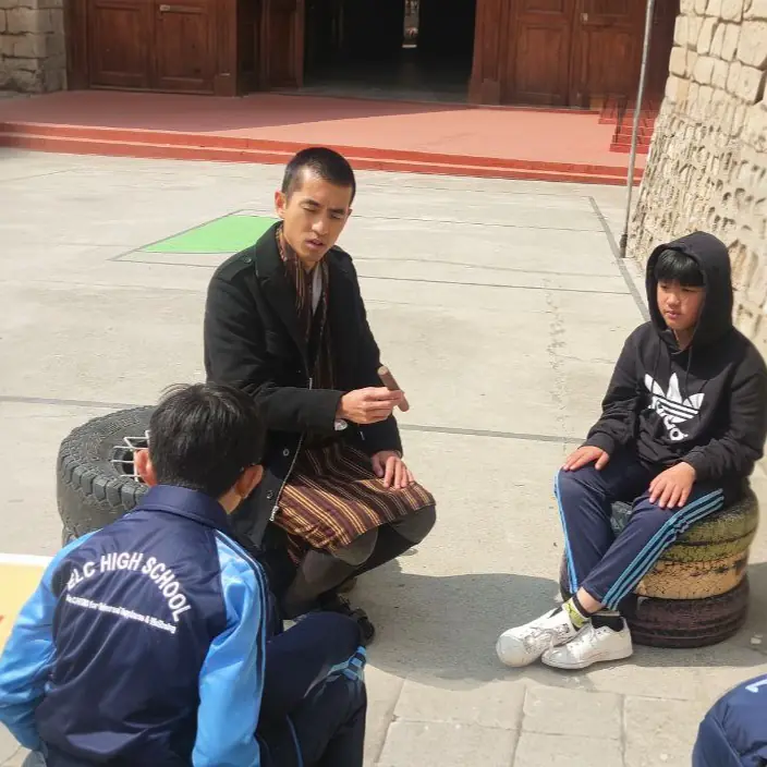 A teacher crouched in the courtyard talking with two students sitting on painted tire seats